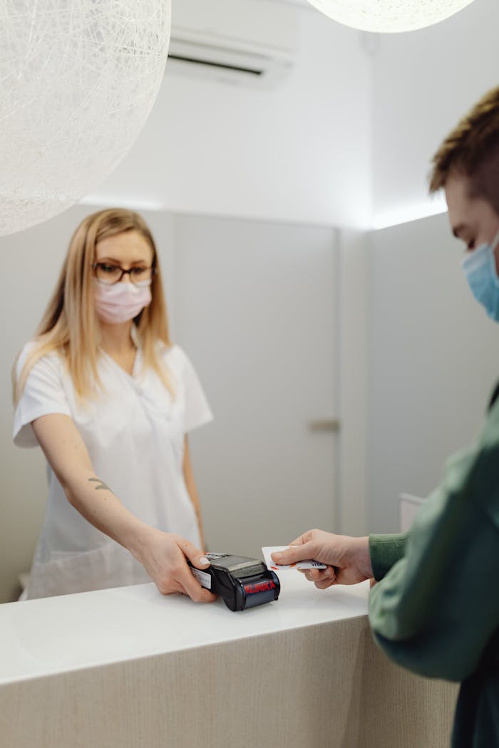 A masked patient pays at a clinic counter, using a card terminal handled by a healthcare professional.